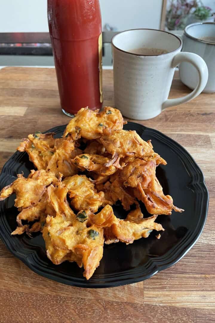 A plate of fried onion pakoda with a bottle of ketchup and two cups of chai.