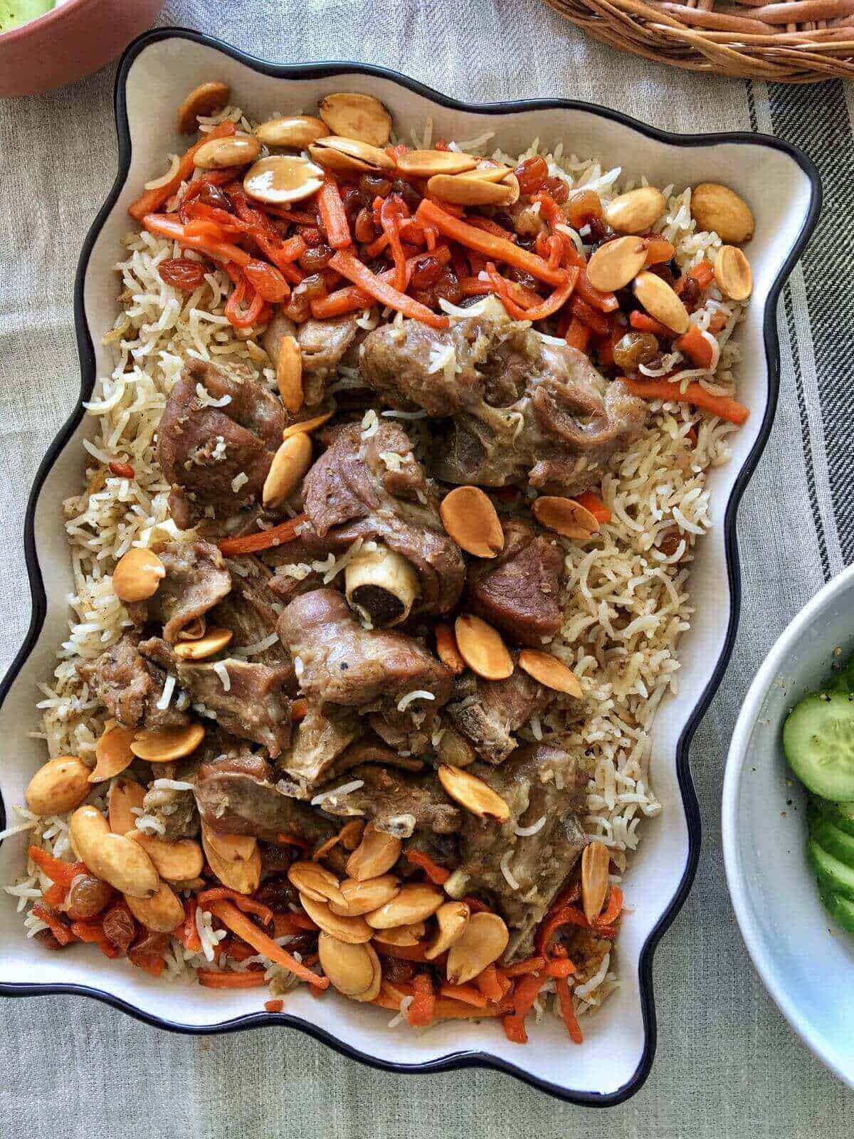 a rectangular serving dish filled with rice and topped with pieces of lamb meat in the centre and caramelized carrots, raisins and almonds along the corners.