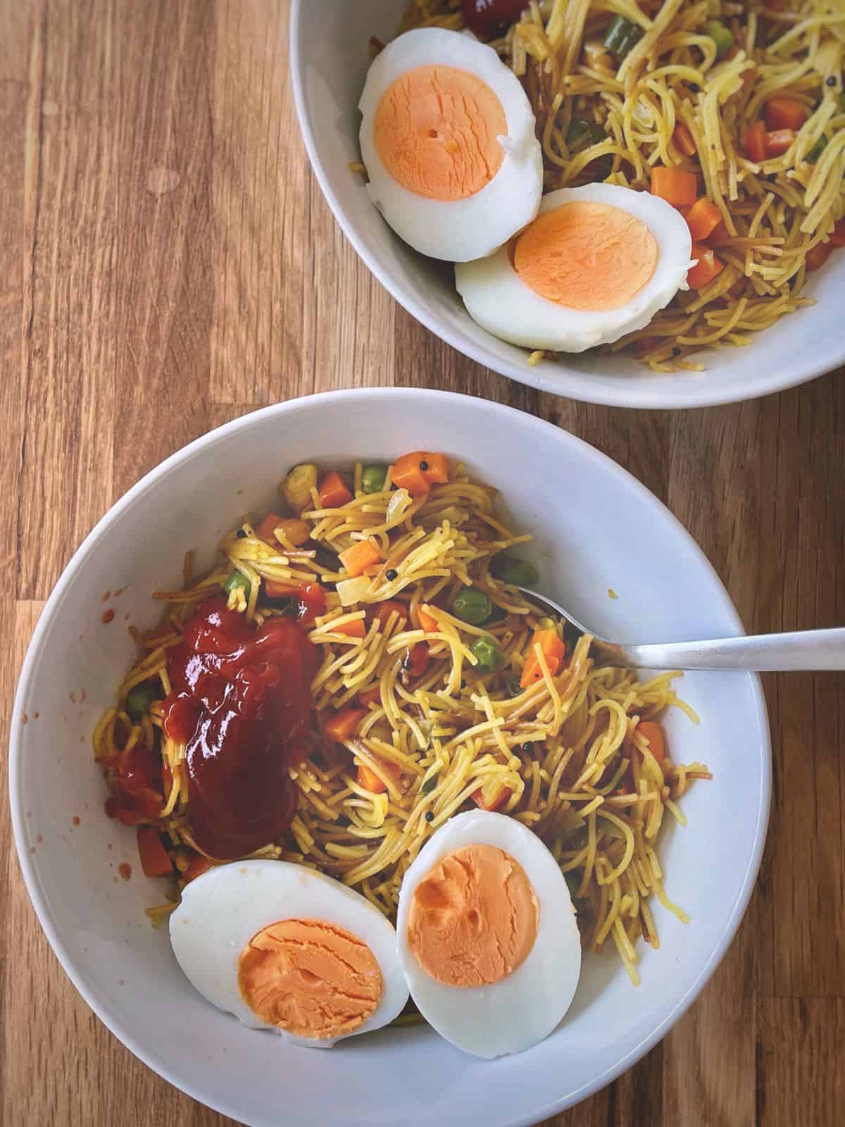 Two bowls of semiya upma topped with some ketchup and a halved boiled egg. 