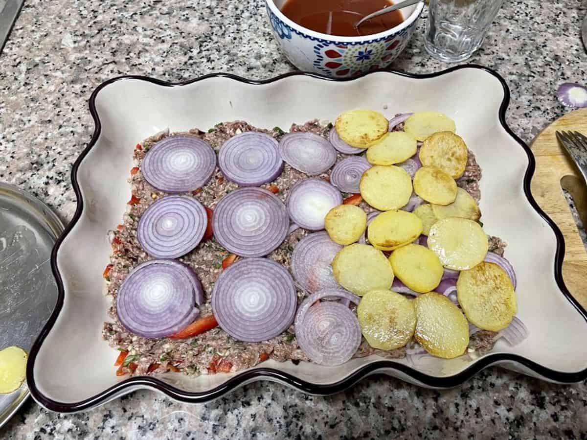 a layer of sliced onion and pan-fried potato slices over kafta 