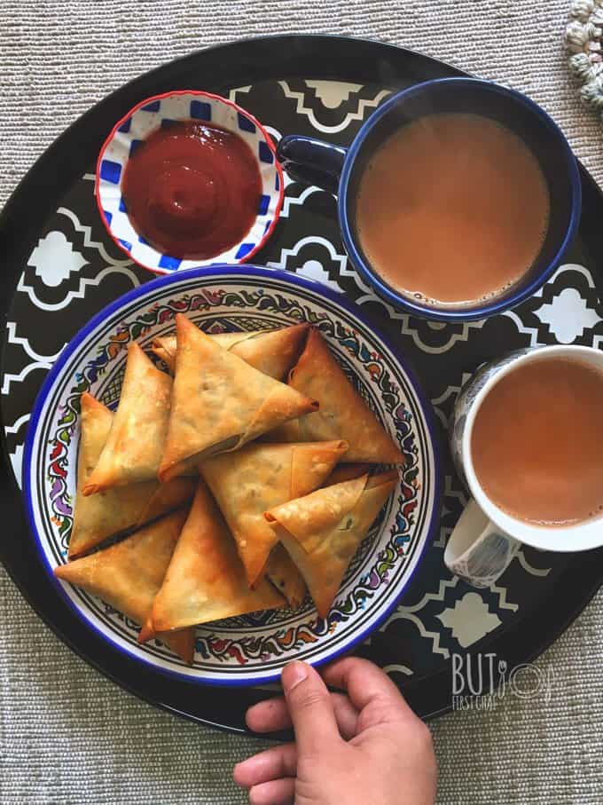holding a tray with a plate of samosas, two cups of chai and a bowl of ketchup