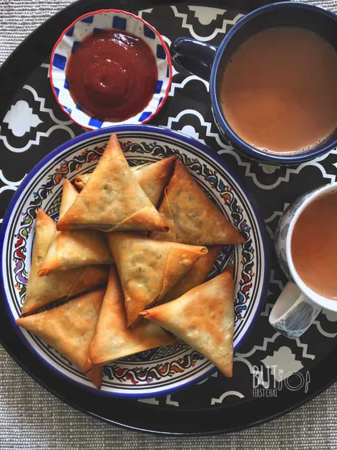 A tray with a plate of baked samosas, two cups of chai and a bowl of ketchup
