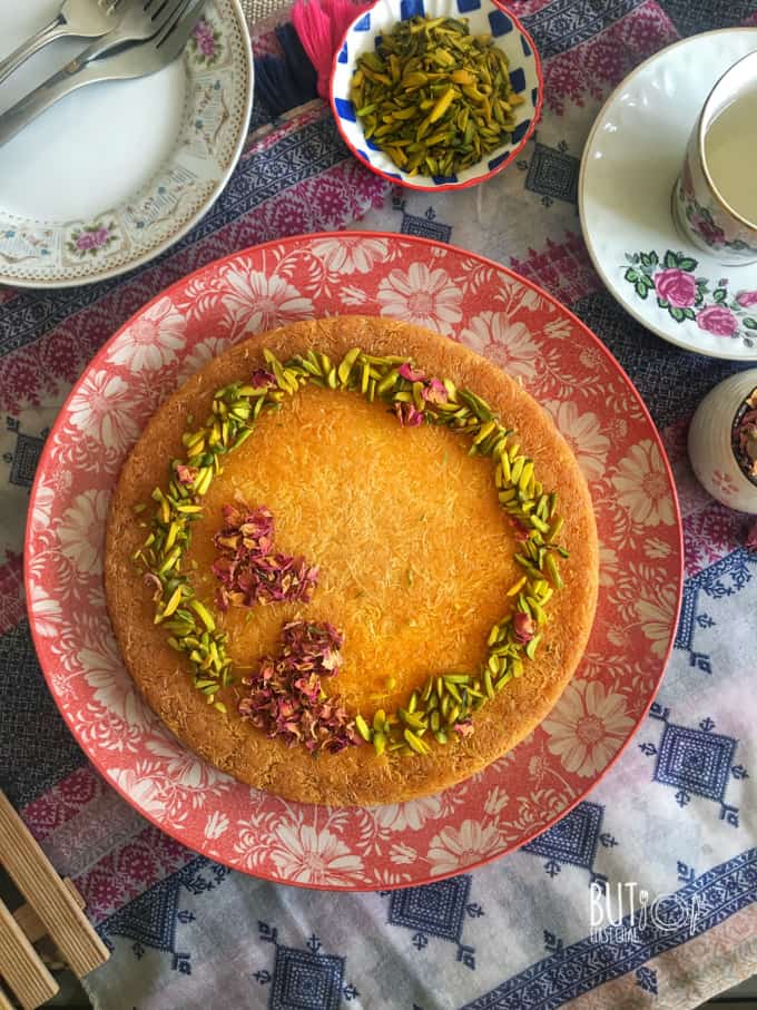 cream kunafa garnished with pistachios and rose petals served along with aromatic syrup and more nuts.