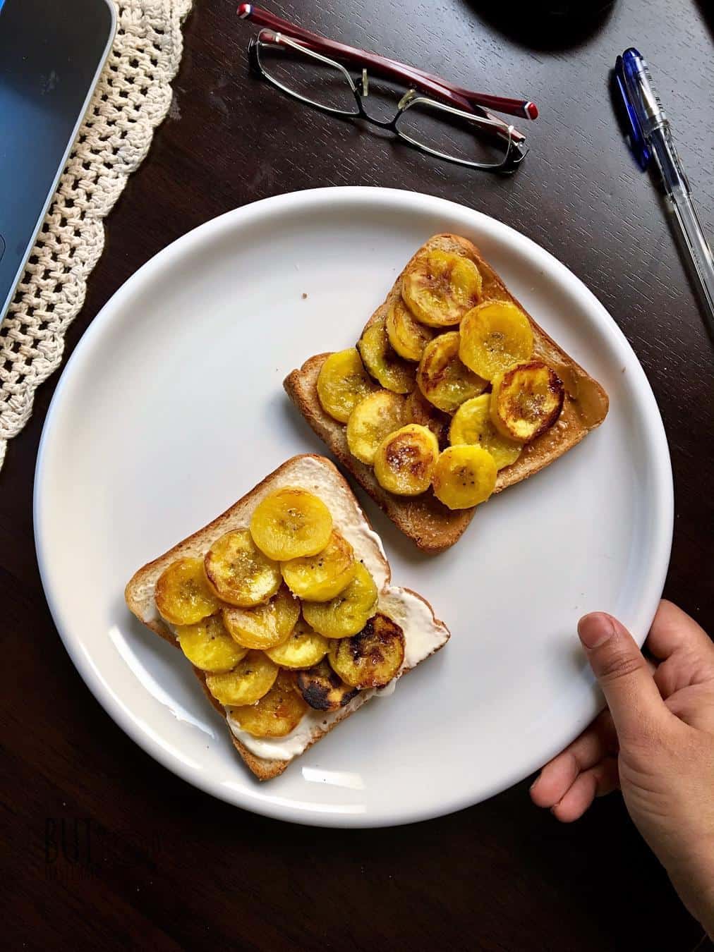 pan roasted ripe plantains neatly arranged over the bed of two toasted sliced bread with labneh. They are placed on a round white plate beside a spectacle and pen.