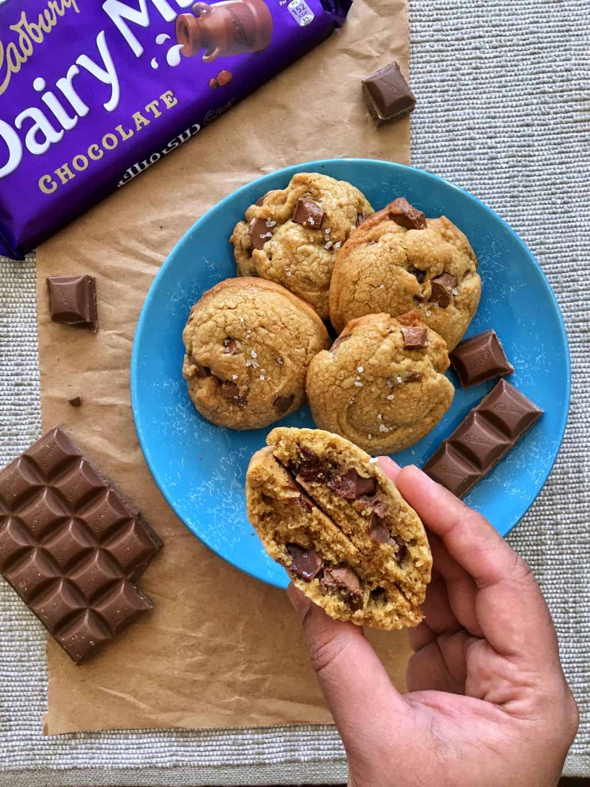 a blue plate with 4 dairy milk cookies with a hand holding a cookie cut to display the chocolate chunks. A few blocks of dairy milk chocolate is scattered along with a dairy milk chocolate wrapper.
