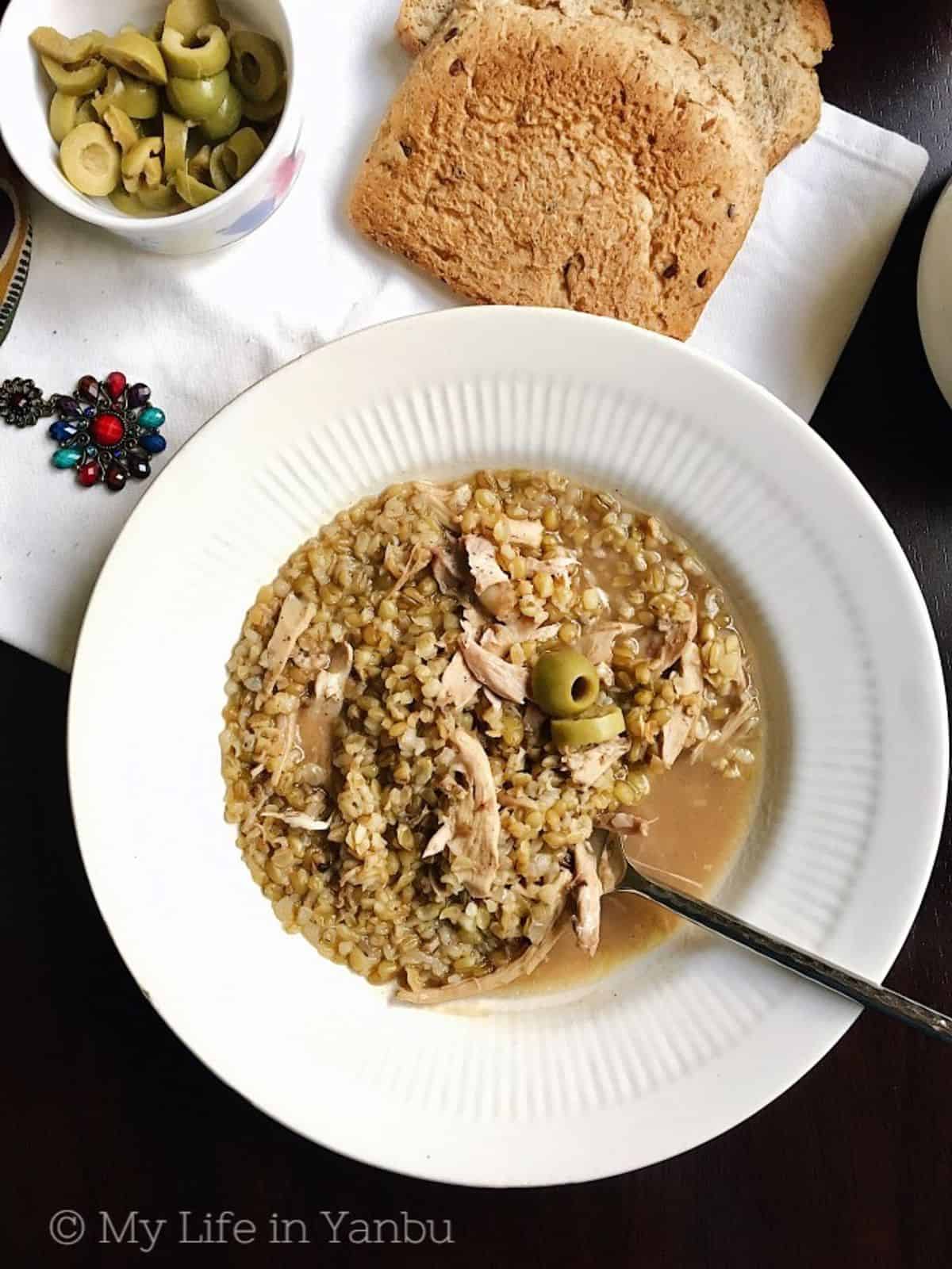 A bowl of Freekeh soup with chicken along with a slice of bread and some olives.