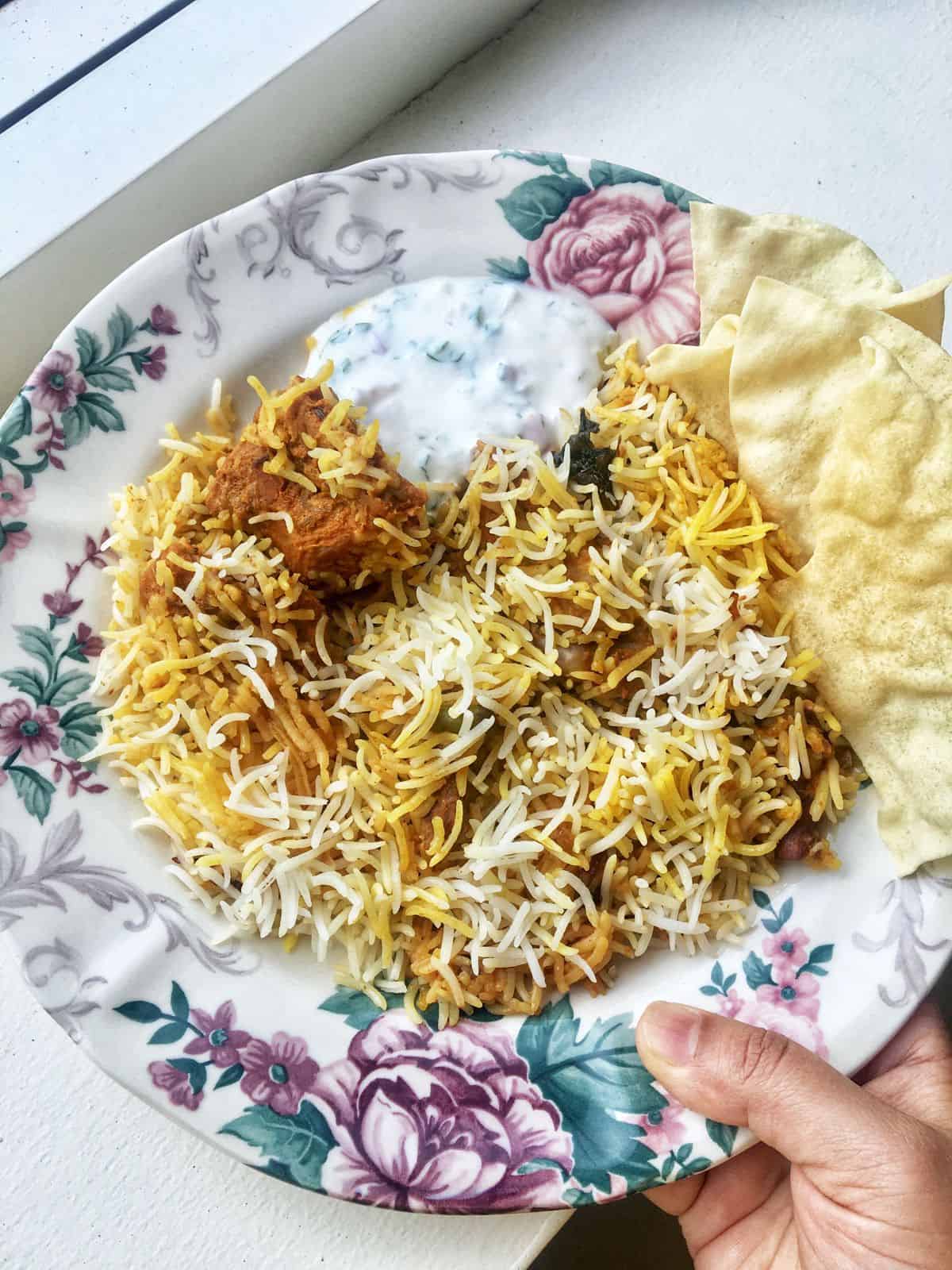 a plate of kacchi mutton biryani served with raita and pappadams in the side