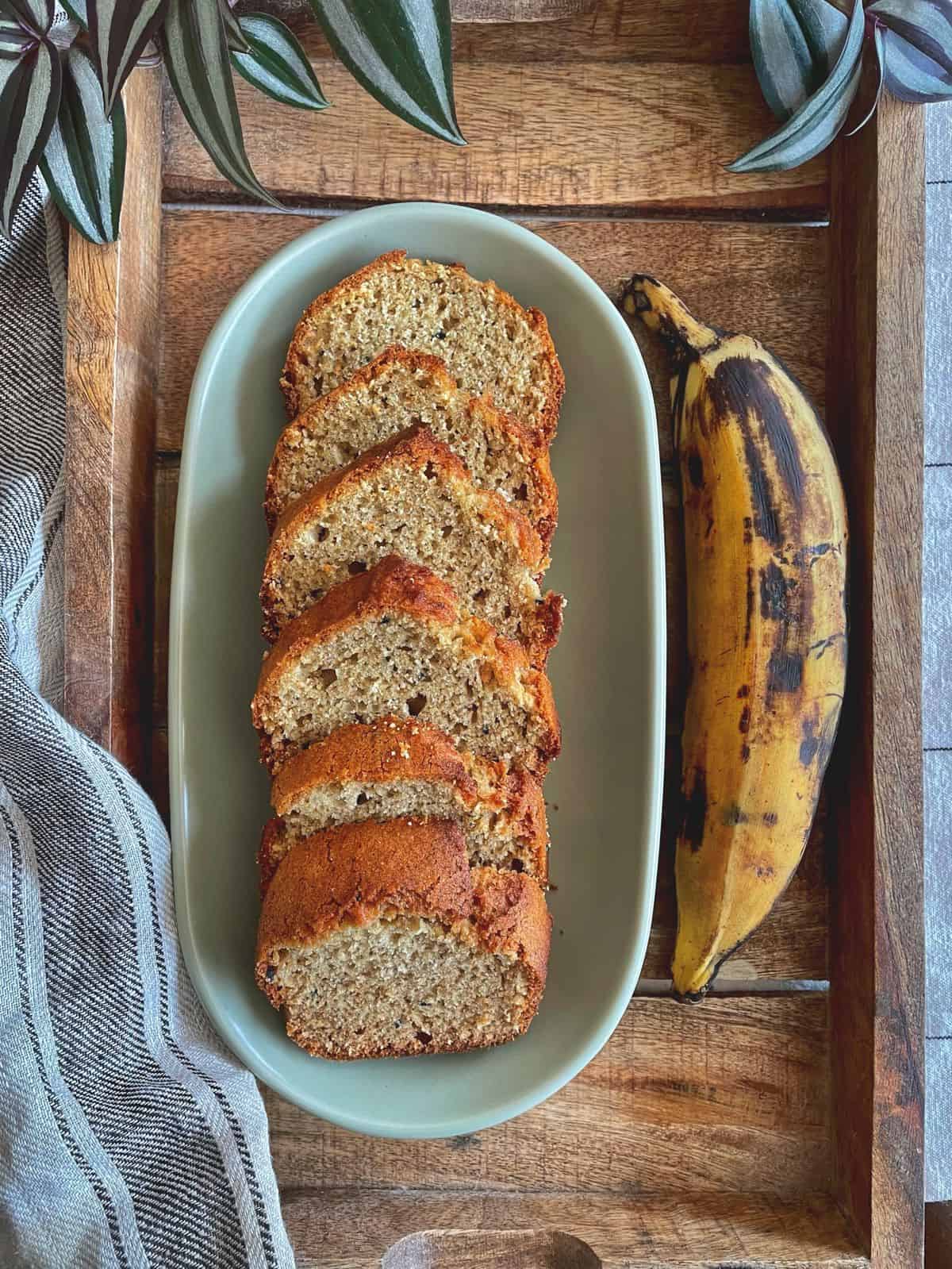 A sliced loaf of plantain bread on an oval plate with a ripe plantain placed beside it.