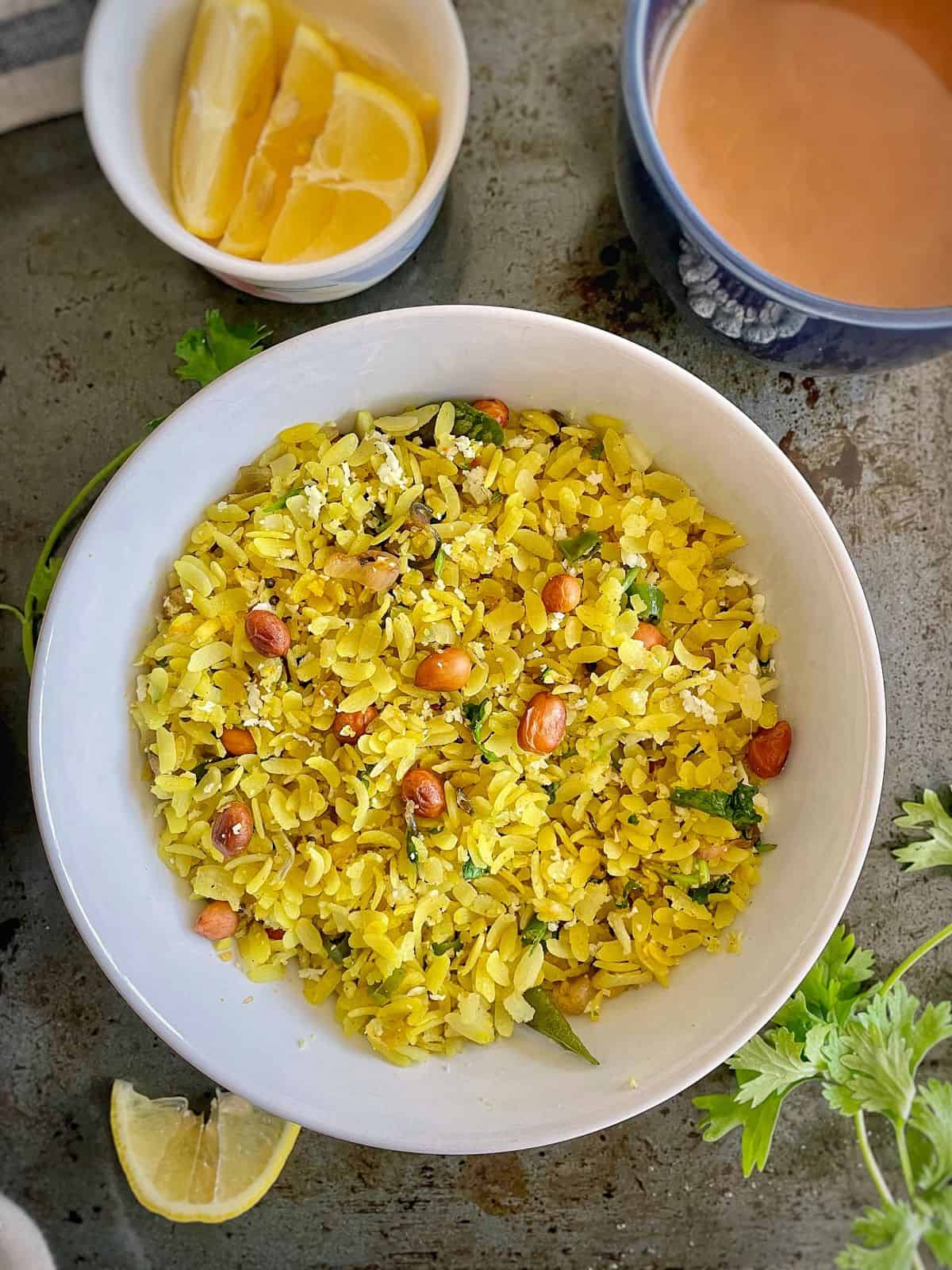 A bowl of kanda poha served with a cup of chai and lemon wedges