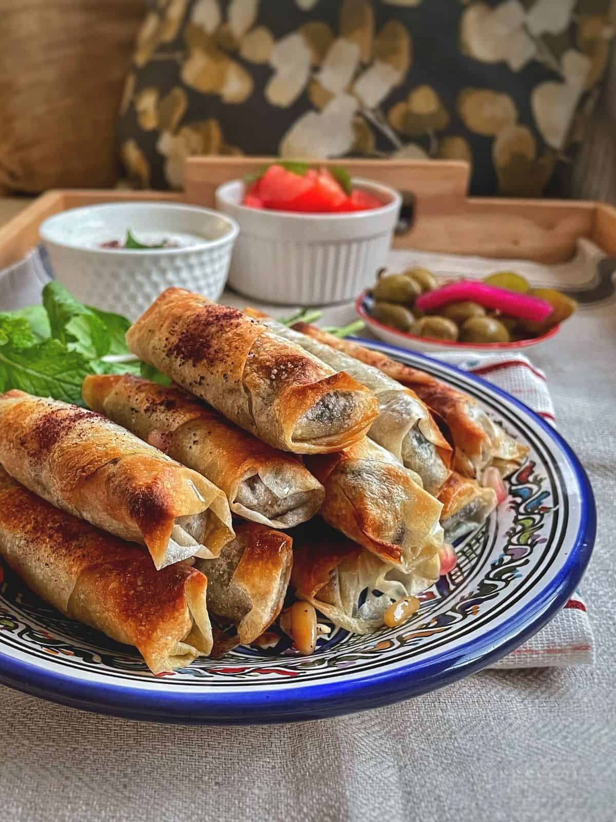 Musakhan rolls stacked on a blue plate with bowls of olives and tomatoes by the side. 