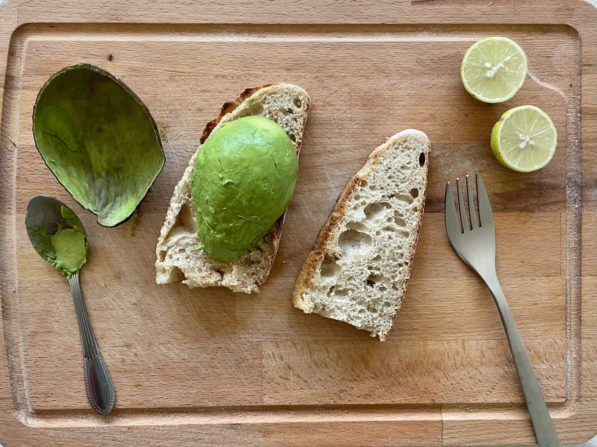 An avocado shell and a spoon used to scoop. A half avocado placed atop the toasted bread. A fork on the cutting board. 