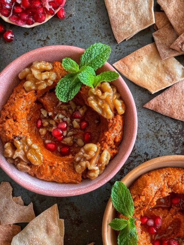 A bowl of roasted capsicum dip garnished with walnuts, pomegranate arils, mint leaf and olive oil.