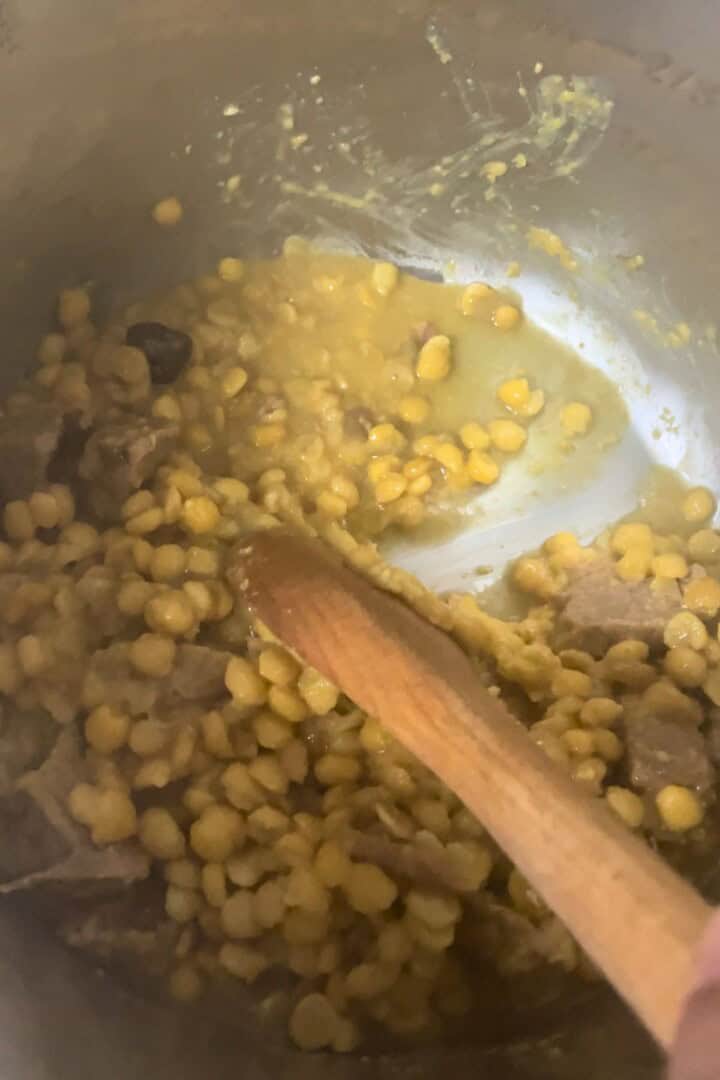 A person stirring a pot of meat and dal with a wooden spoon.