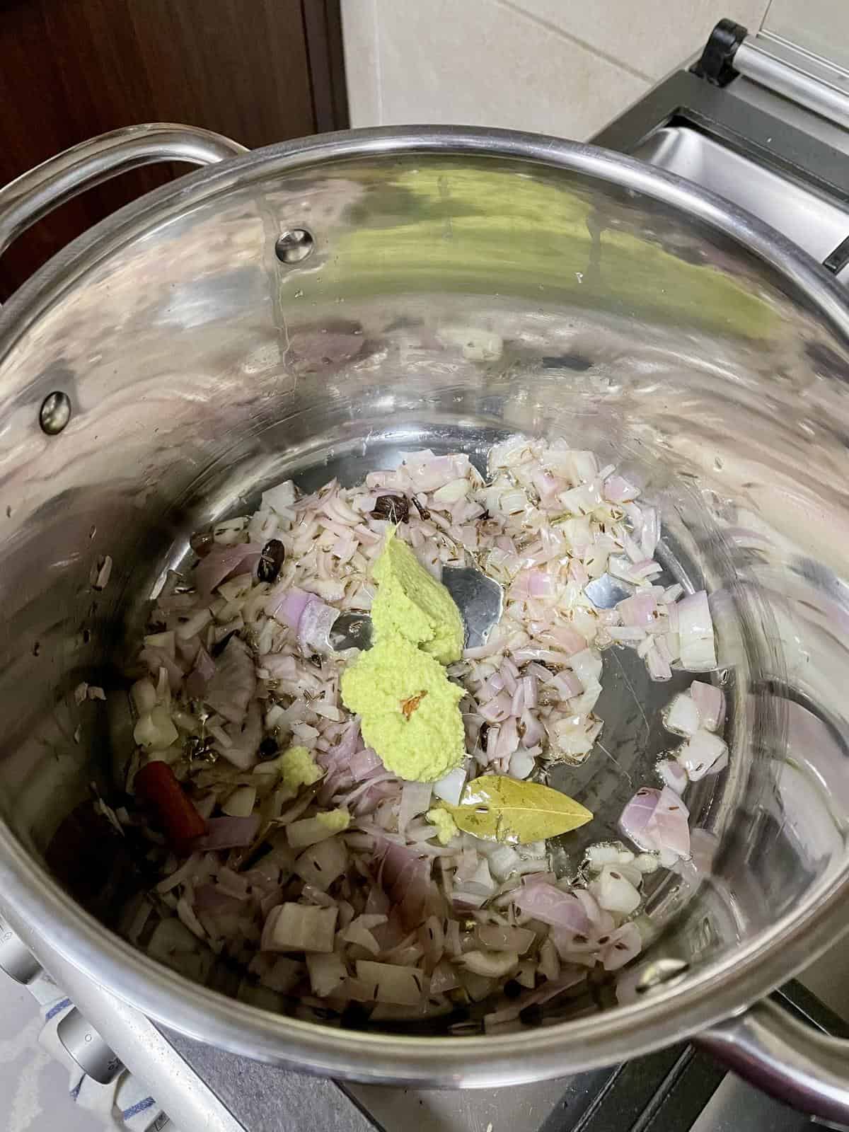 Chopped onions, garlic, bay leaf, and green chili in a stainless steel pot for cooking biryani.