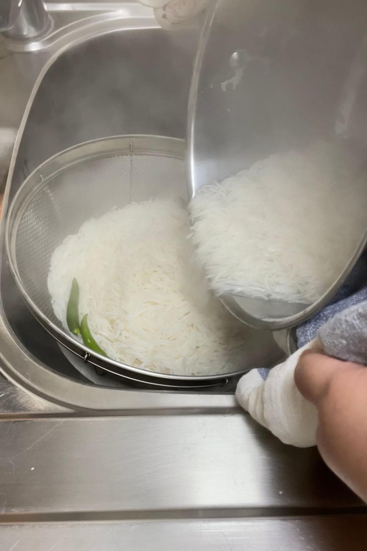 Steamed par-boiled basmati rice with green chilies in a stainless steel pot being drained into a colander. 