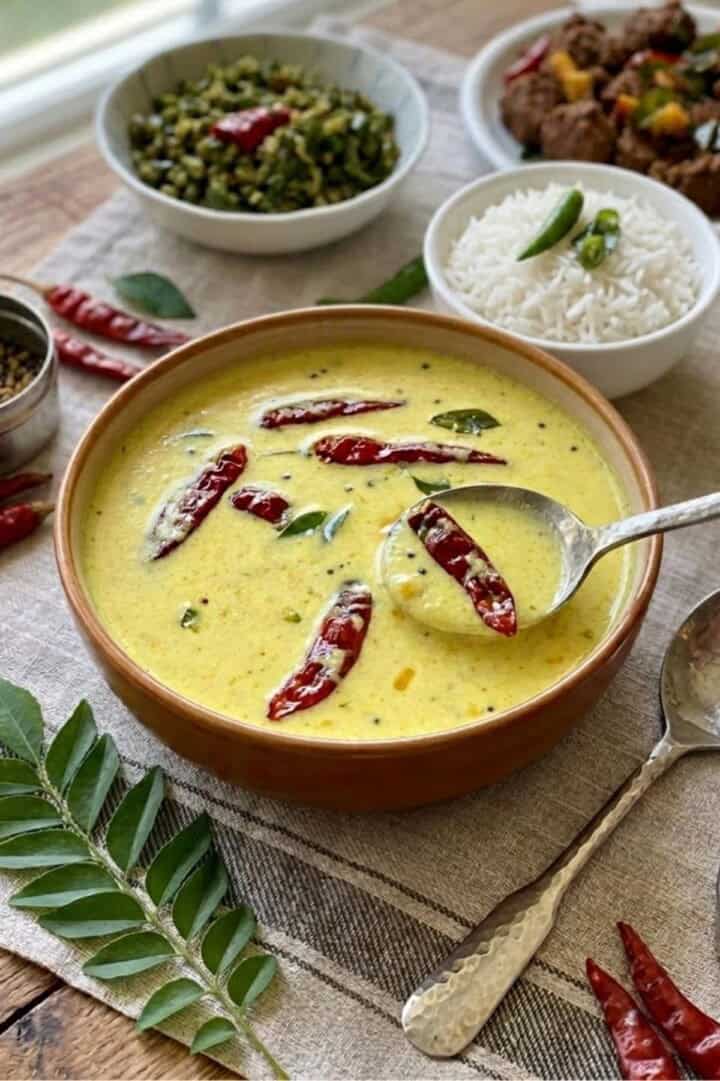 A bowl of moru curry with a spoon served on the table along with rice, thoran and beef fry.